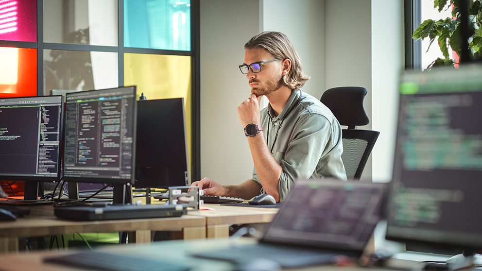 Mann mit Brille denkt konzentriert vor Bildschirmen mit Code – Symbolbild für kritisches Denken im KI-Zeitalter