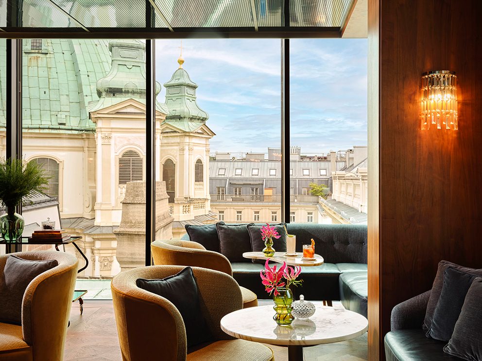Rosewood Vienna: Ein eleganter Raum mit kleinen Tischen, Sofas und Sesseln mit Blick auf ein altes Gebäude in Wien.