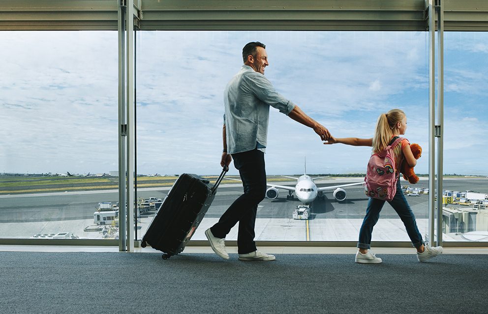 Ein Vater und seine kleine Tochter gehen über einen Flughafen, im Hintergrund sieht man das wartende Flugzeug als Symbolbild für Reiseversicherungen