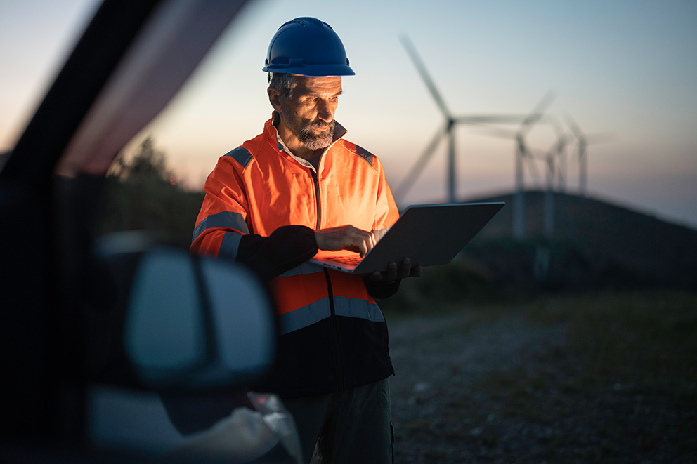 Ein Mann in Arbeitskleidung und Schutzhelm steht vor einem Windpark und arbeitet am Laptop, als Symbol für das Thema KI im Mittelstand