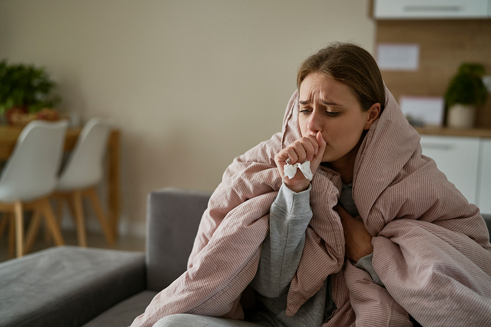 Kranke Frau sitzt hustend in eine Decke gehüllt auf dem Sofa – Symbolbild zur Attestpflicht